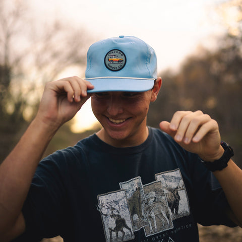 Person wearing a blue cap with a logo and a black t-shirt with graphic designs outdoors.