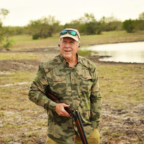 Man in camouflage clothing holding a rifle in an outdoor setting with a pond and trees in the background.