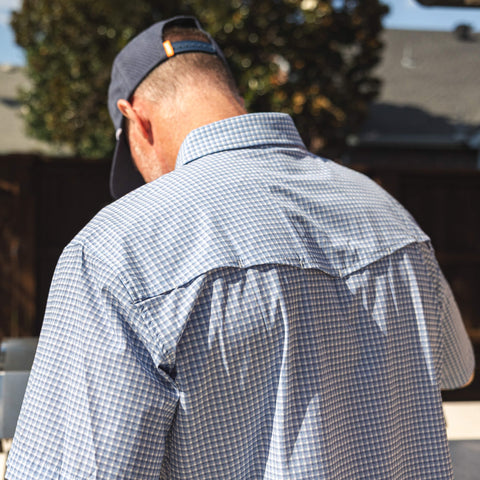 Man wearing a blue checkered shirt and cap, standing outdoors with blurred background