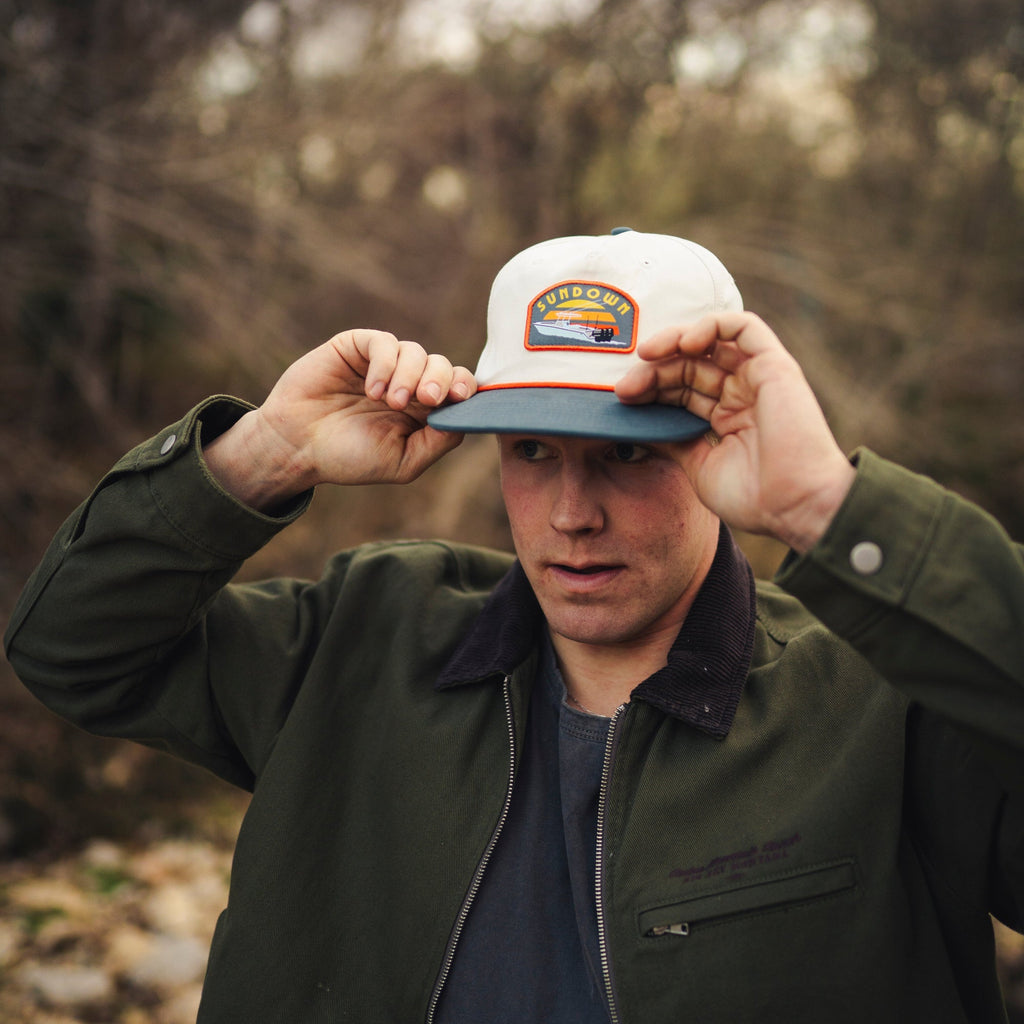 Man adjusting a cap with a patch boat logo in the woods