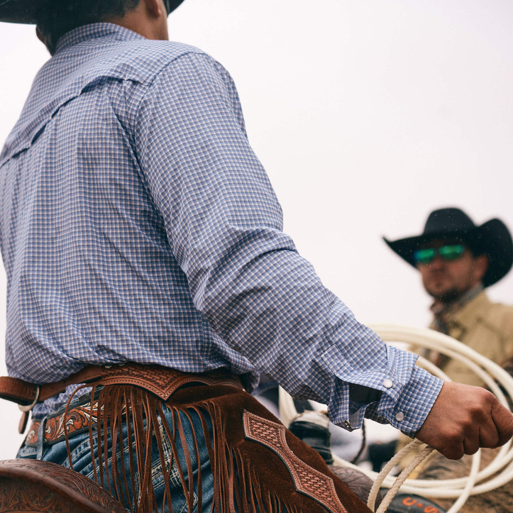 Cowboy in checkered shirt and chaps holding a lasso with another cowboy in the background.