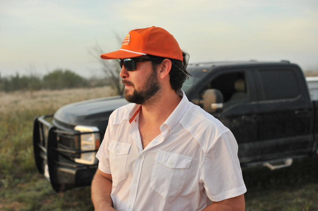side view of man standing in front of truck wearing white performance field shirt short sleeve and blaze sundown hat