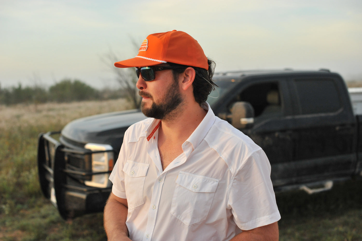 side view of man standing in front of truck wearing white performance field shirt short sleeve and blaze sundown hat