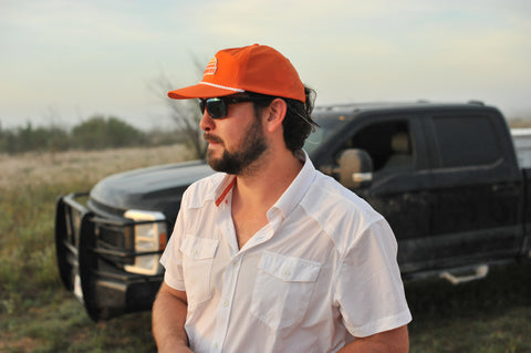 side view of man standing in front of truck wearing white performance field shirt short sleeve and blaze sundown hat