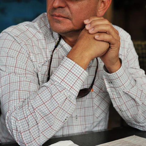 Man wearing a checkered shirt with his hands clasped, sitting at a table.
