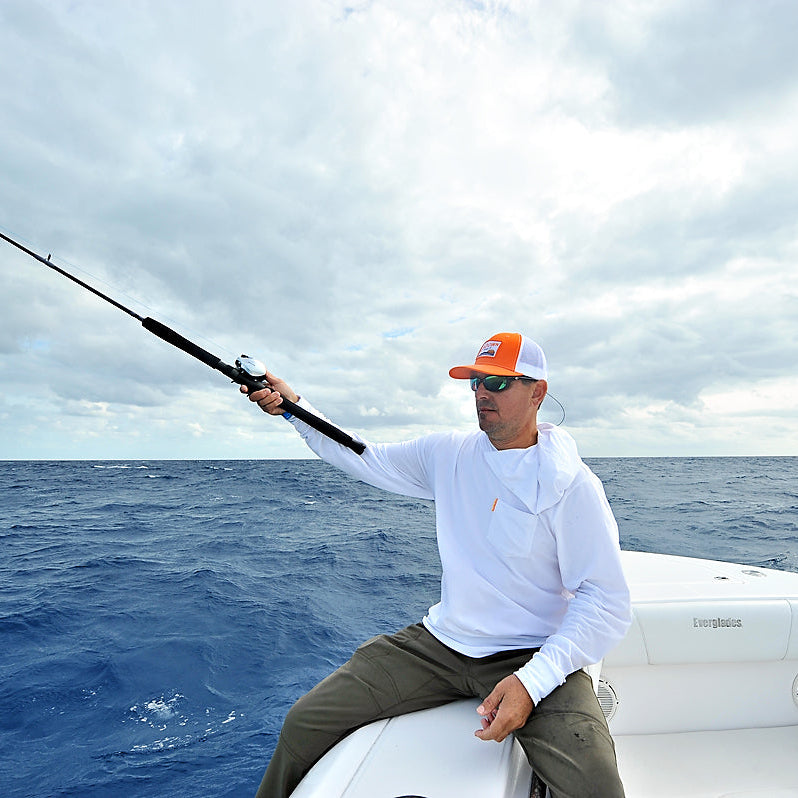 A man wearing a white long-sleeve shirt, olive pants, and an orange and white cap sits on the edge of a white boat, holding a fishing rod over the blue ocean under a cloudy sky.