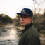 Person wearing a blue cap with branding, standing outdoors near a river.