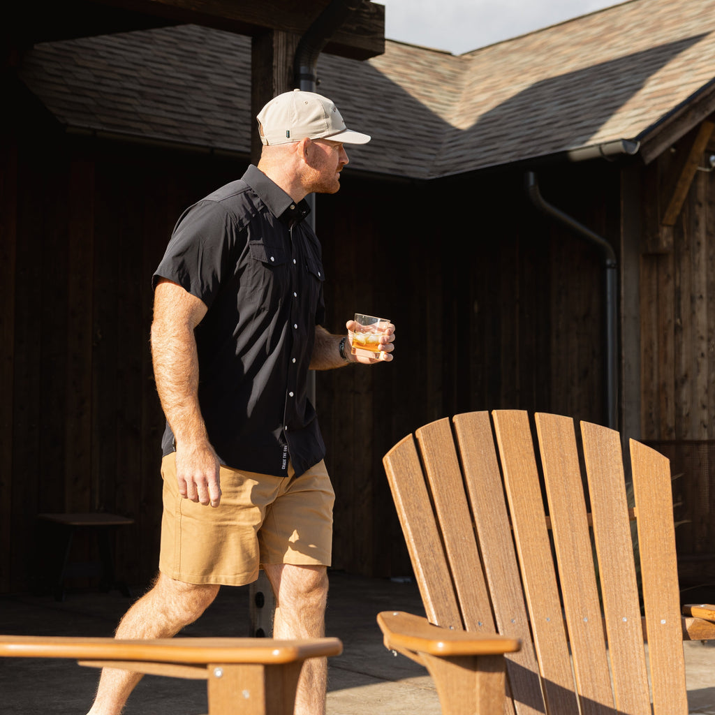 A man wearing a black short-sleeve button-up shirt, khaki shorts, and a beige baseball cap walks across a wooden deck holding a drink, with Adirondack chairs and a rustic cabin in the background.