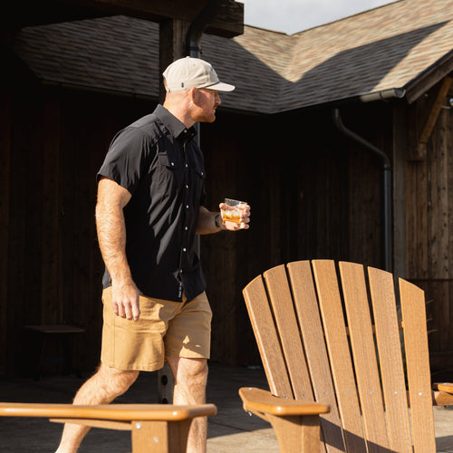 A man wearing a black short-sleeve button-up shirt, khaki shorts, and a beige baseball cap walks across a wooden deck holding a drink, with Adirondack chairs and a rustic cabin in the background.