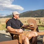A man sitting in a wooden Adirondack chair outdoors, holding a drink and looking to the side, with mountains, a grassy field, and a blue sky with clouds in the background.