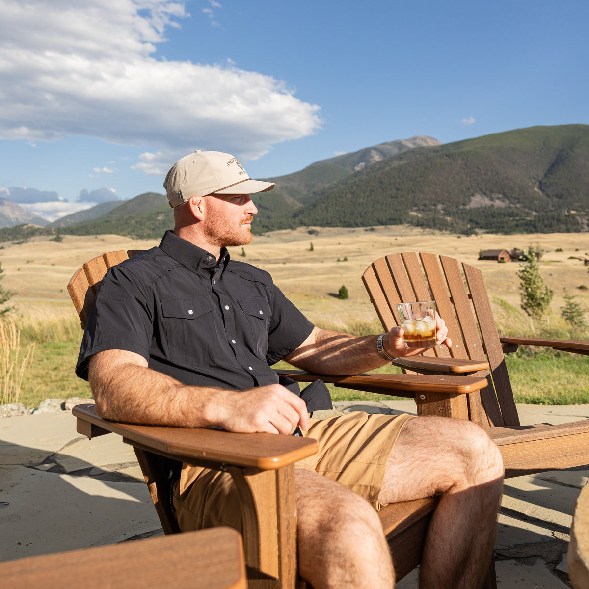 A man sitting in a wooden Adirondack chair outdoors, holding a drink and looking to the side, with mountains, a grassy field, and a blue sky with clouds in the background.