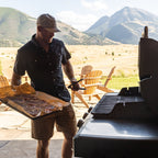 A man wearing a cap and dark shirt stands outdoors near a grill, holding a tray of food in a backyard setting with mountains and chairs in the background.